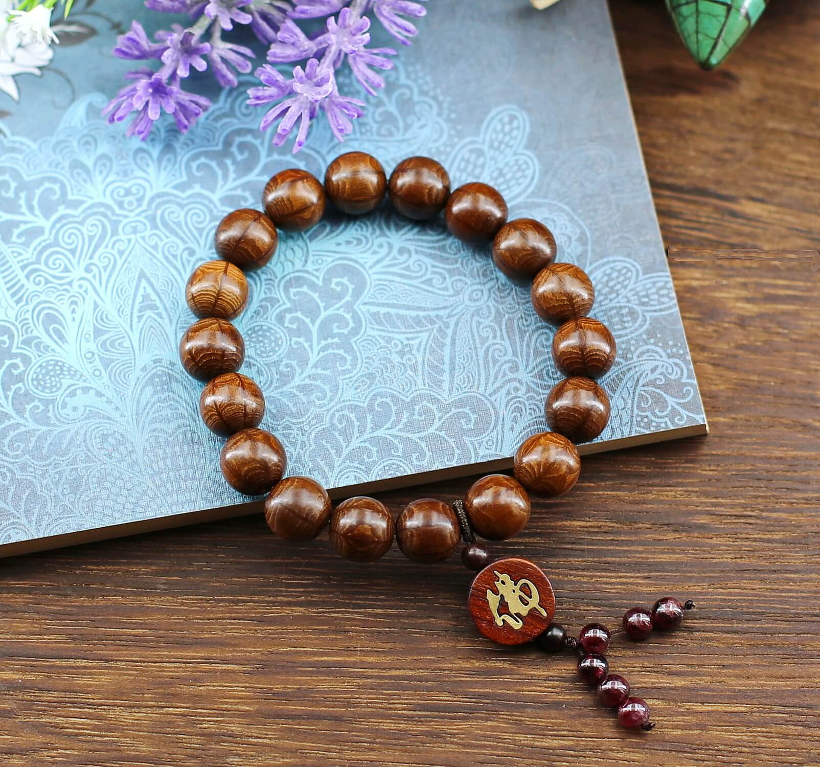 Ce bracelet-mala tout en raffinements est composé de perles de bois d'Abélia foncé. Il est accompagné d'un pendant de bois de Santal rouge incrusté à l'or biface du nom du Bouddha et orné de six perlettes du Grenat: un bel ensemble pour un bracelet-chapel